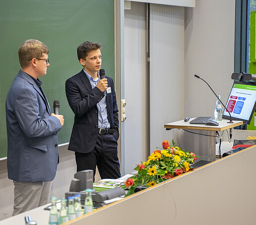 Two young scientists explain their project on a green board.