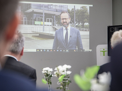 Videoübertragung über Beamer an einer Wand mit Staatsminister Gemkow. Gäste schauen sich seine Rede an.