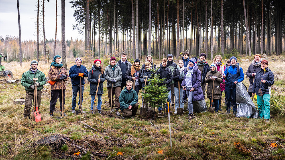 A group of students and university management in the Zittau city forest with Wiedekopfhauen stand in a row for a group picture. A planted fir tree stands in the middle.