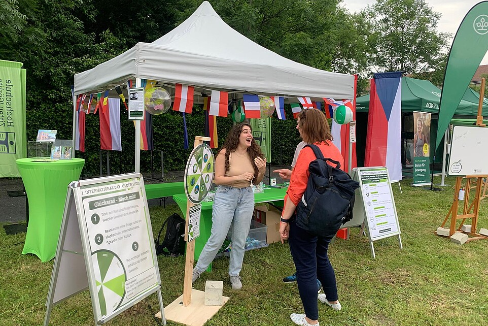 Festival participants chat in front of the International Day Pavilion. In front of it is a wheel of fortune. International flags hang from the pavilion.
