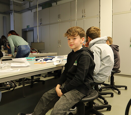 A student sits at a table in the materials lab and smiles at the camera.
