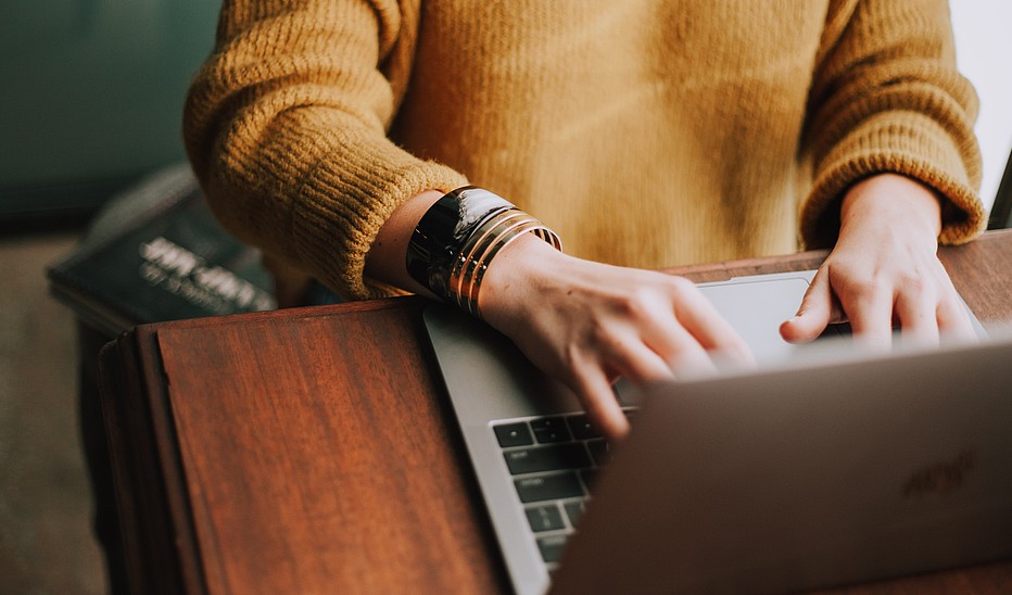 Photo by Christin Hume on Unsplash Eine Person sitzt an einem Laptop. Man sieht nur ihren gelben Pullover und ihre Hände, die auf der Tastatur schreiben.