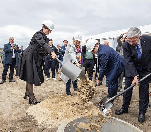 The rector and the other project participants each throw a shovelful of sand into a hole in the ground. Guests of the event stand in the background.