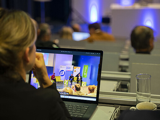 In the lecture hall, a participant is sitting in a seat, looking forward and has a laptop open in front of her.