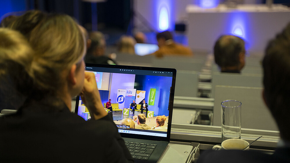 In the lecture hall, a participant is sitting in a seat, looking forward and has a laptop open in front of her.