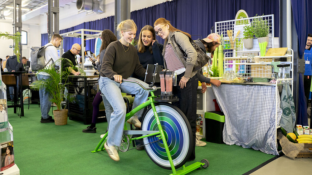 A prospective student cycles on the smoothie bike, two companions stand around her.