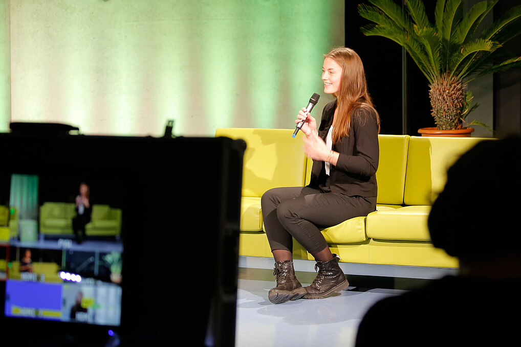 A student sits on a couch in a studio and holds a microphone in her hand.