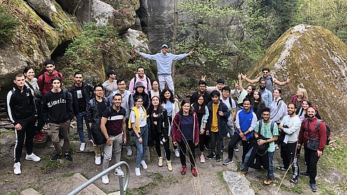 Eine Gruppe internationaler Studierender steht vor einer Gesteinsformation des Berges Oybin und lächelt in die Kamera.