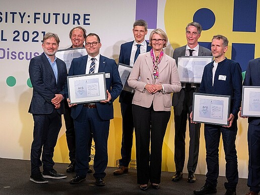 Auf einem Podium stehen sieben Männer, mit einer Urkunde in der Hand, in der Mitte steht die Bundesbildungsministerin und lächelt in die Kamera.