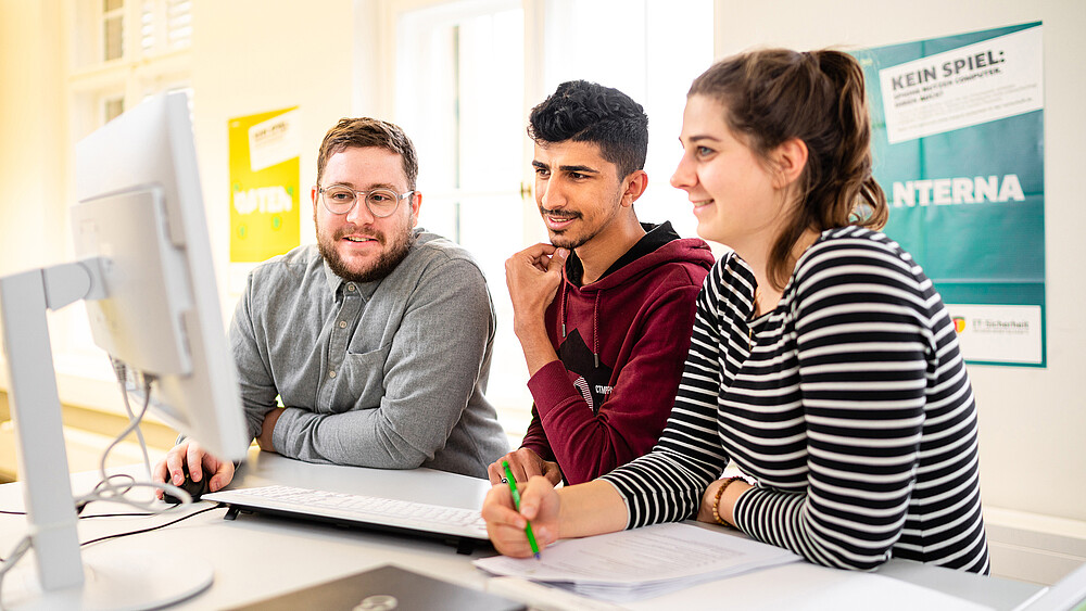 Three students are standing at a bar table, smiling and looking at a laptop in front of them.