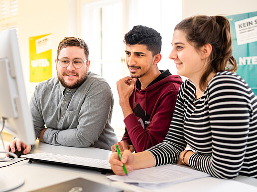 Three students are standing at a bar table, smiling and looking at a laptop in front of them.