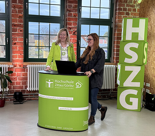 A contributor and the presenter stand behind an HSZG counter in the studio. On the left in the foreground is a black camera. Four HSZG cubes are piled up to the right of the counter.