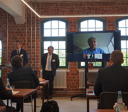In a lecture hall, speakers stand in front of their audience and a screen is set up next to them, on which another speaker is connected.