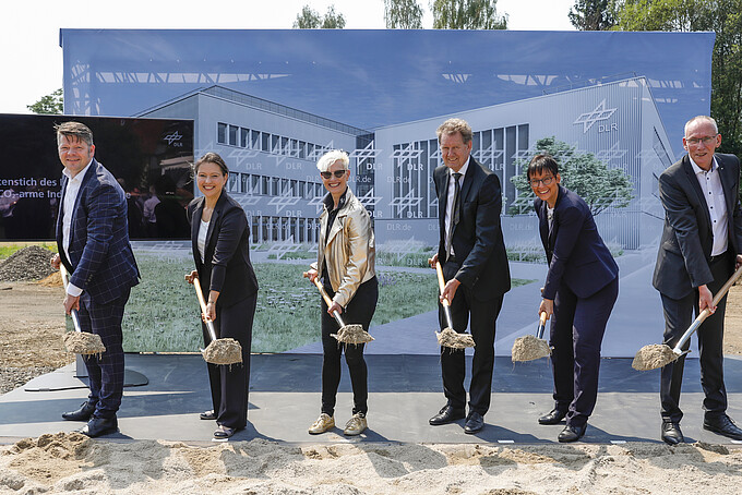 Initiators of the DLR Institute each hold a spade filled with soil in front of them and look into the camera. A press wall is set up in the background, on which the design of the future institute is depicted.