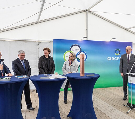 The participants in the CircEcon project stand in a white tent at blue bar tables and talk in front of a blue and green press wall with the CircEcon logo printed on it.