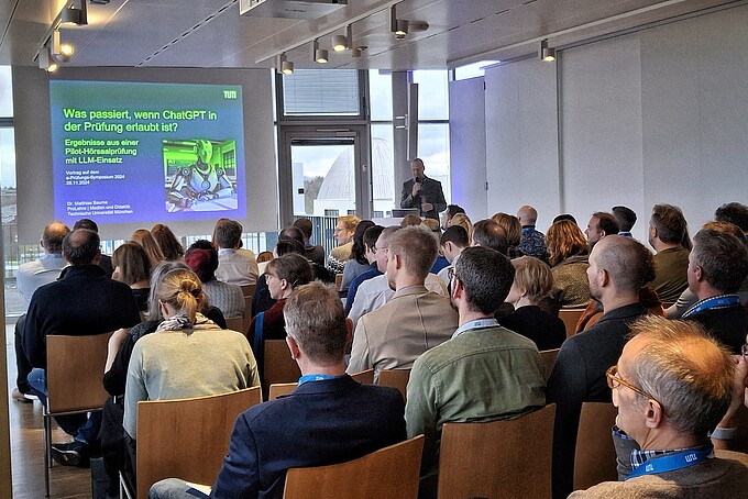 In a seminar room, a participant gives a speech next to a projector presentation in front of a full row of chairs.