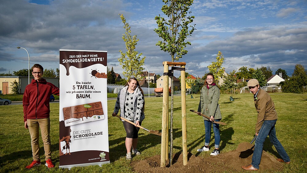 Four people plant a tree. Next to it is a roll-up with the advertising message.