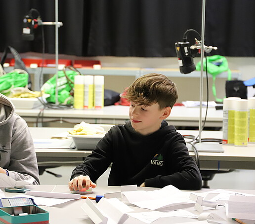 A student sits at the table and looks at the handicrafts of the person sitting next to him.