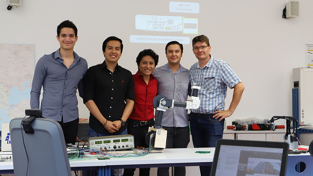 Four Mexican students and a university employee stand behind a table with a robotic arm on it.