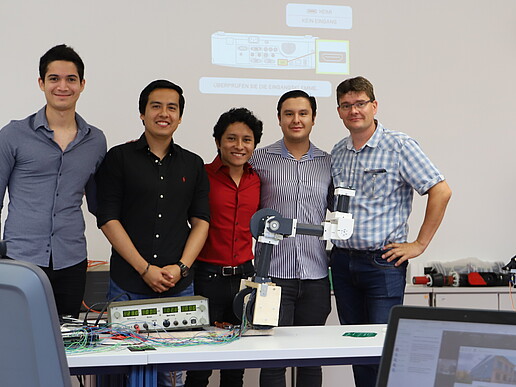 Four Mexican students and a university employee stand behind a table with a robotic arm on it.