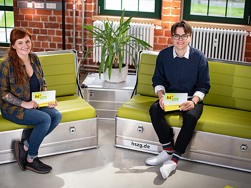 A female presenter and a young male presenter are sitting on two separate sofas, smiling at the camera and holding presentation cards in their hands.