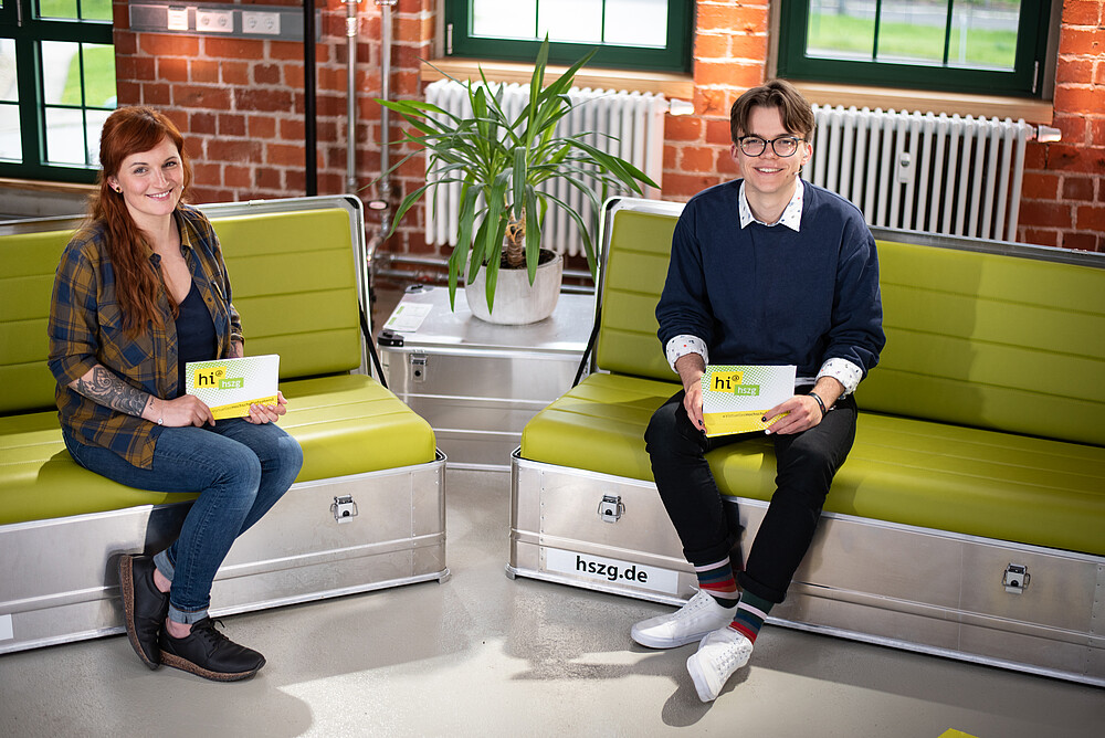A female presenter and a young male presenter are sitting on two separate sofas, smiling at the camera and holding presentation cards in their hands.