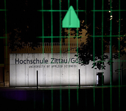 The Zittau/Görlitz University of Applied Sciences lettering shines brightly, with a green illuminated construction fence in the foreground.