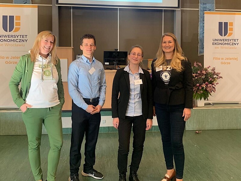 Four representatives of the HSZG stand next to each other in a hall decorated with roll-ups and smile into the camera.