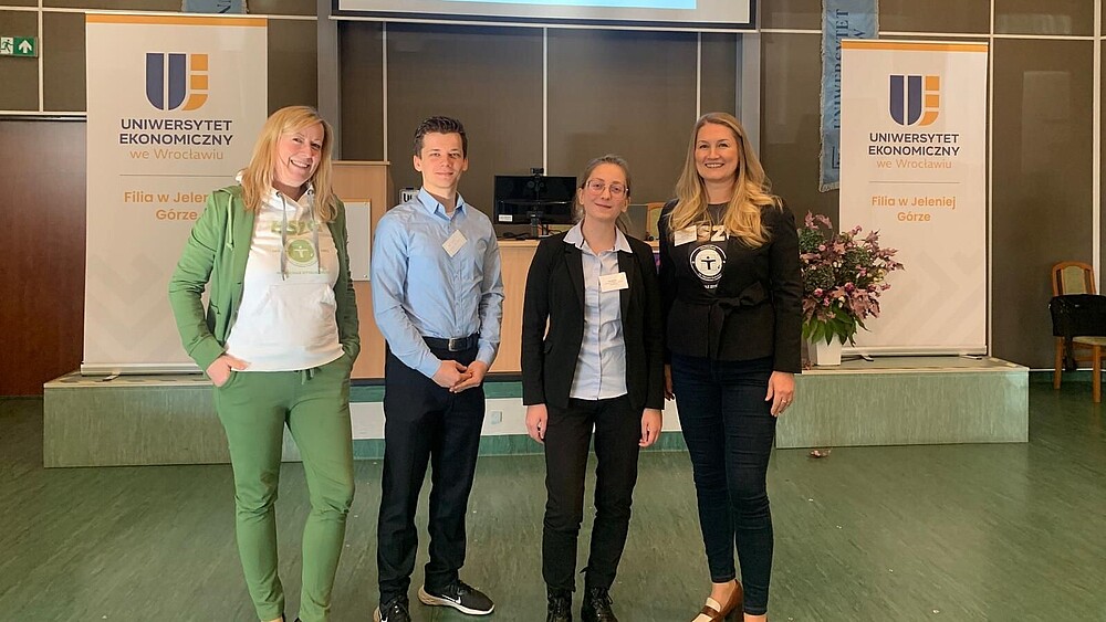 Four representatives of the HSZG stand next to each other in a hall decorated with roll-ups and smile into the camera.