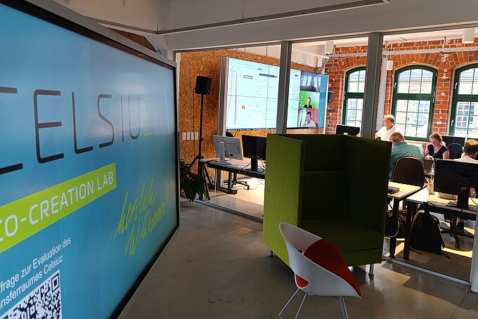 The Celsiuz logo on a screen stands in the foreground of a workroom where people are sitting at tables and looking at a large screen.