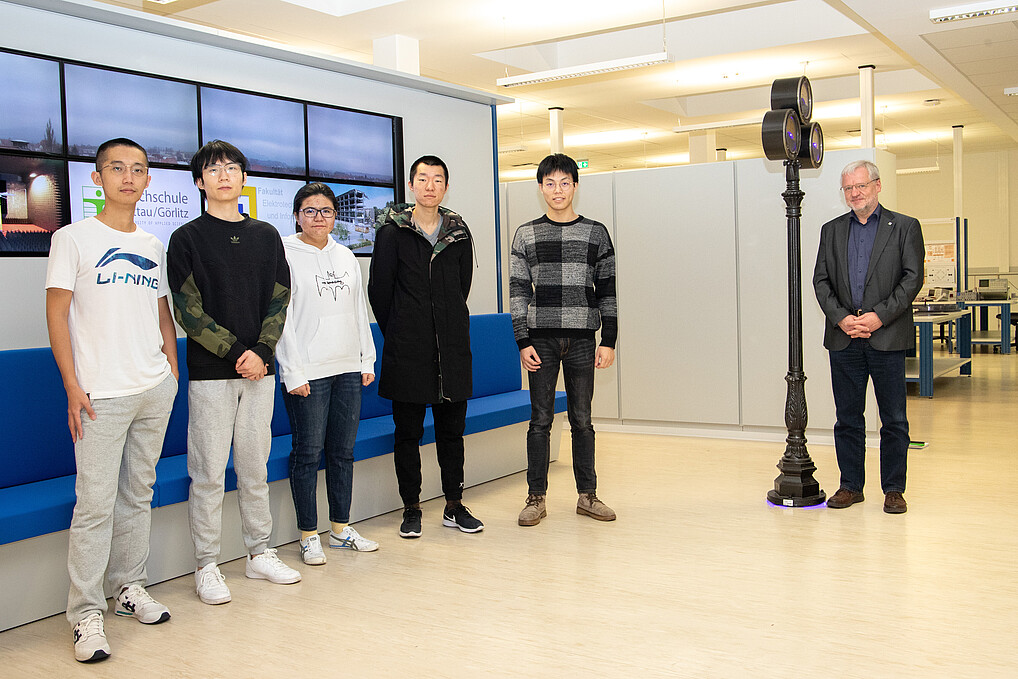 A group of five Chinese students and Professor Frank Worlitz stand in front of a large LED screen in a laboratory hall.
