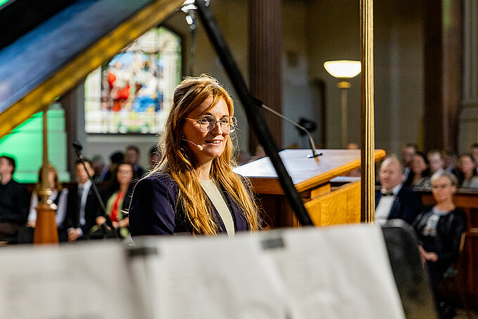 Victoria Hohlfeld stands next to the grand piano, singing, and looks toward the audience.