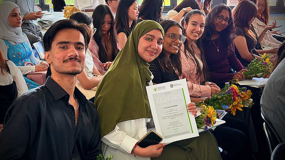 Students from the preparatory college at Zittau/Görlitz University of Applied Sciences are seated in Building Z IV during the graduation ceremony. In the foreground, a graduate proudly holds her diploma, while other graduates sit in their seats with flowers and certificates.