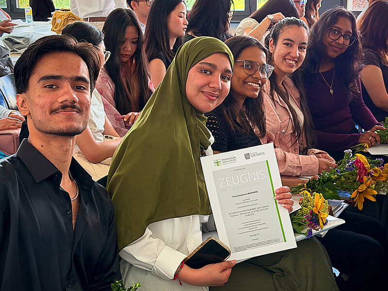 Students from the preparatory college at Zittau/Görlitz University of Applied Sciences are seated in Building Z IV during the graduation ceremony. In the foreground, a graduate proudly holds her diploma, while other graduates sit in their seats with flowers and certificates.