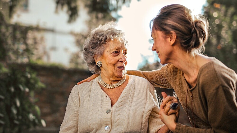 A young woman puts her arm over the shoulder of an older lady and takes her by the hand. They smile at each other.