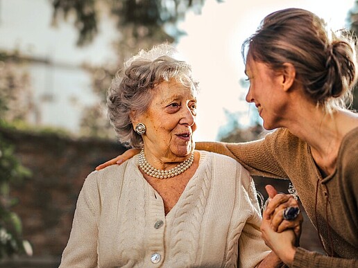 A young woman puts her arm over the shoulder of an older lady and takes her by the hand. They smile at each other.