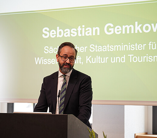 A man in a suit with glasses is speaking at a lectern. Behind him is a presentation slide with his name and his function as Minister of Science.