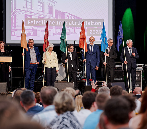 Dekane und Dekanin halten ihre Fakultätsfahne auf der Bühne.