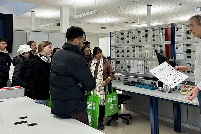 Dr. Wolfgang Menzel erklärt den Besucher in einem weiträumigen Labor der Fakultät Elektrotechnik und Informatik der HSZG verschiedene Aspekte der technischen Ausstattung. Das Labor ist modern eingerichtet, mit zahlreichen technischen Tafeln und Geräten, während die Gruppe aufmerksam zuhört.