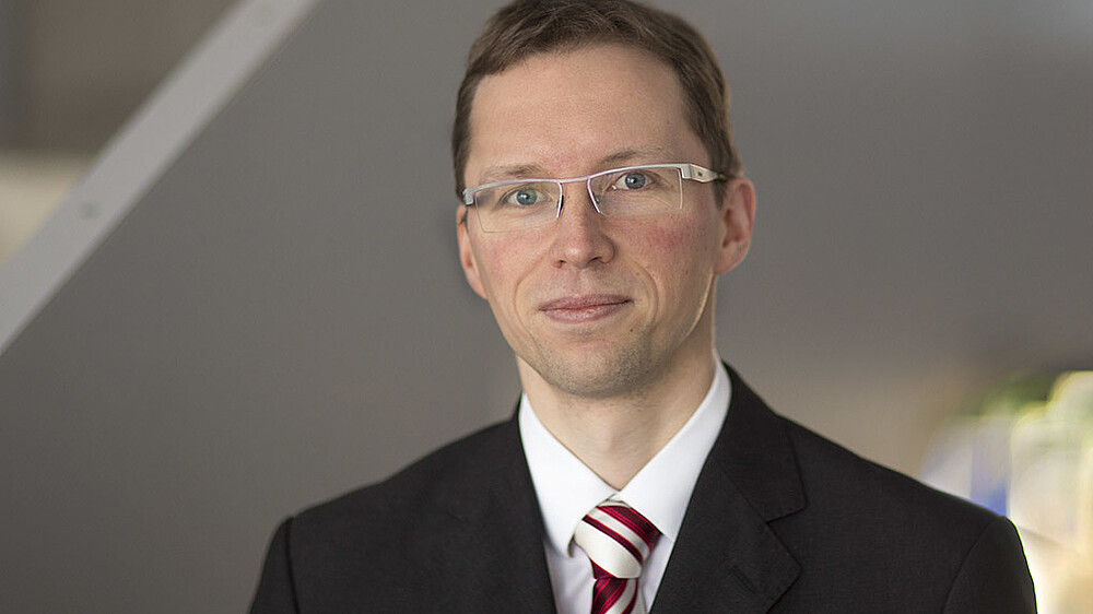 Stefan Kornhuber in a suit with tie and glasses smiles into the camera.