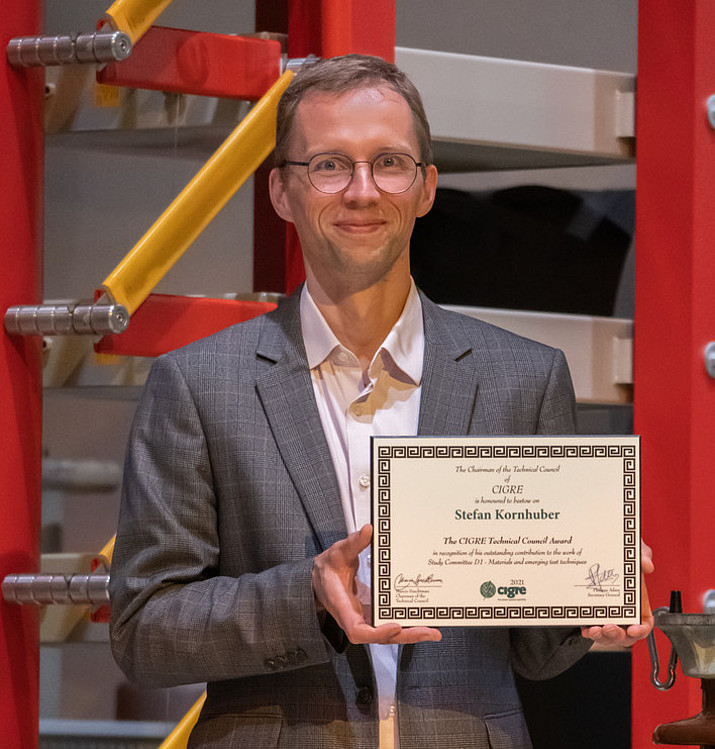 Prof. Kornhuber holds his CIGRE Award certificate up to the camera. He is standing in the high-voltage hall of the HSZG.