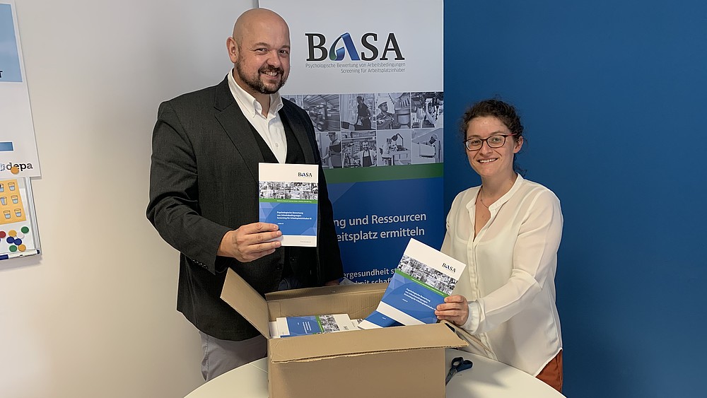 Prof. Matthias Schmidt and BASA employee Katharina Roitzsch hold the new BASA information materials up to the camera and smile.