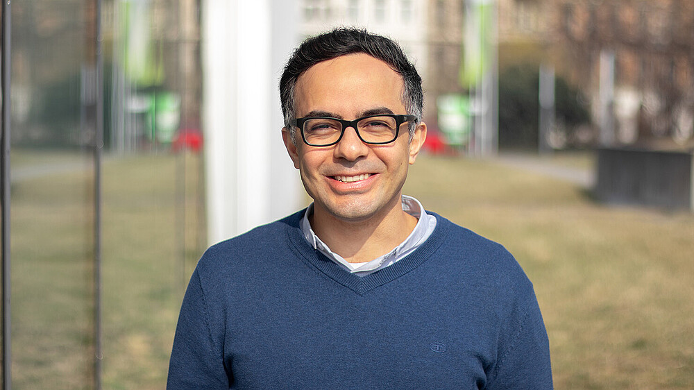 Prof. Massoudi, wearing a blue sweater and glasses, stands outside next to a glass façade and smiles at the camera.
