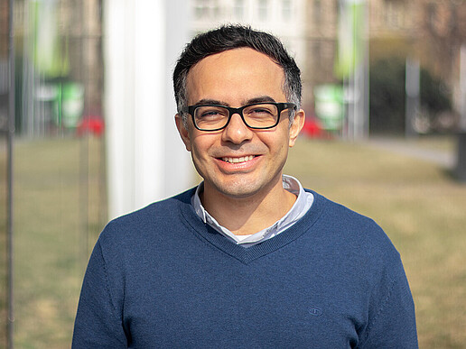 Prof. Massoudi, wearing a blue sweater and glasses, stands outside next to a glass façade and smiles at the camera.