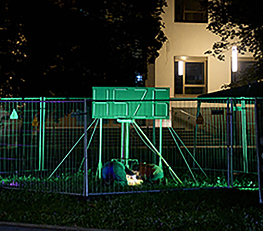 A glowing green light installation in the shape of the HSZG logo stands on the lawn in front of the HSZG building, surrounded by construction fences.