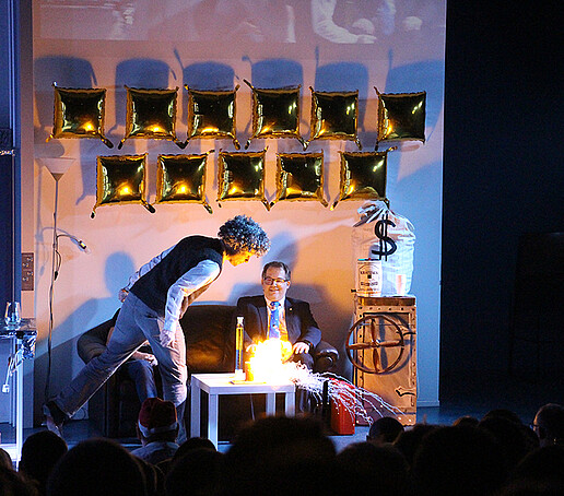 A fire experiment on stage is observed by the principal, who is sitting on the couch.
