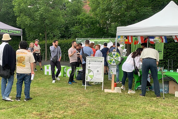 Festival participants chat in front of the International Day Pavilion. In front of it is a wheel of fortune. International flags hang from the pavilion.