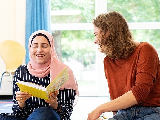 Zwei junge Frauen sitzen in einem Seminarsaal auf dem Boden, die linke Frau hält ein aufgeschlagenes Buch in der Hand. Beide lachen.