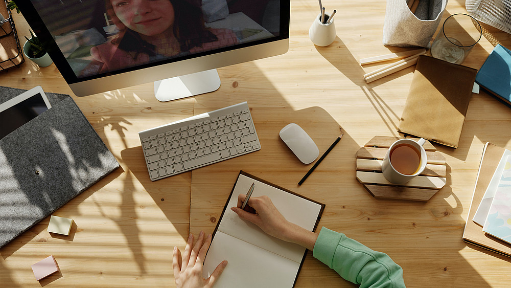 Tisch mit PC, Maus, Tastatur, Kaffeetasse, Stiften und Person die im Notizbuch schreibt. Nebenbei läuft Videokonferenz auf dem PC.