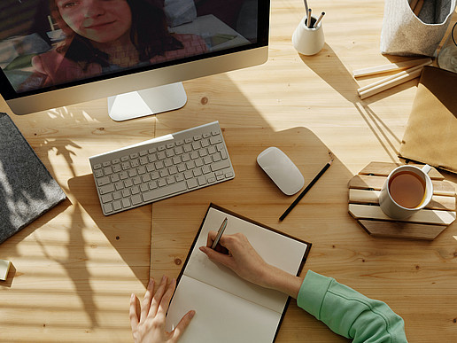 Table with PC, mouse, keyboard, coffee cup, pens and a person writing in a notebook. A video conference is also running on the PC.